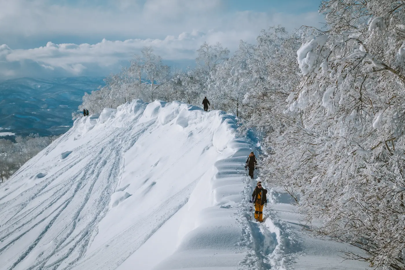 Rider in fila su una cresta di neve, con le montagne e le nuvole sotto