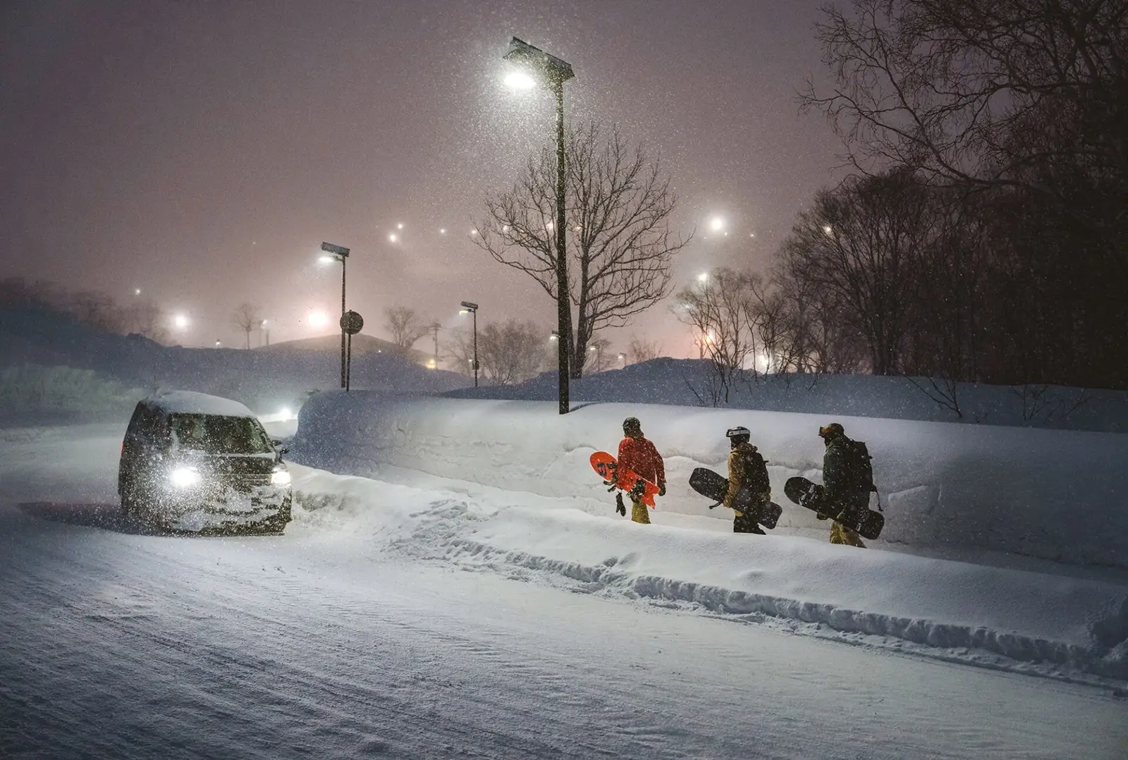 Tre rider con le tavole sotto braccio camminano di notte lungo un muro di neve, il van li segue